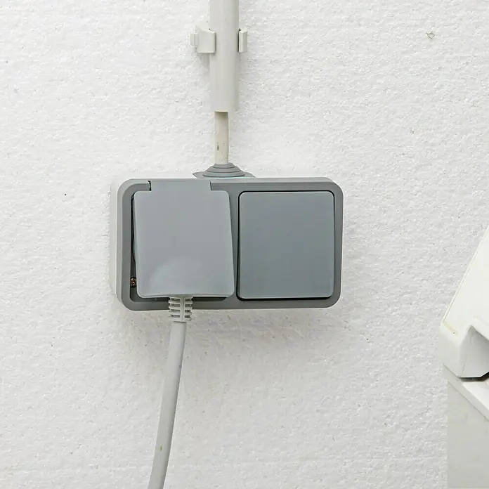 Voltomat Blueline Feuchtraum-Schalterkombination - Grau, Ausrichtung Schalterprogramm: Horizontal, Aufputzmontage, IP54 - Eine graue Doppelsteckdose mit Schutzklappen an einer weißen Wand, ein Stromkabel ist eingesteckt.