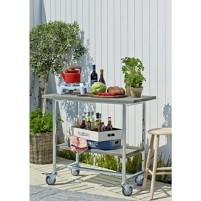Plus Gartentisch / Pflanztisch Urban - 100 x 50 x 90 cm, Graubraun, 1 Boden - Ein grauer Servierwagen aus Metall mit Speisen und Getränken darauf, vor einem weißen Holzzaun im Freien.