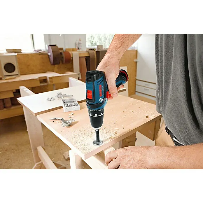 Bosch Professional Taladro atornillador percutor de batería GSB 12V-15 - 12 V, 2 baterías, 2 Ah, 30 Nm, 2 baterías y cargador - Primer plano de un taladro inalámbrico Bosch en uso, haciendo un agujero en una tabla de madera en un taller.