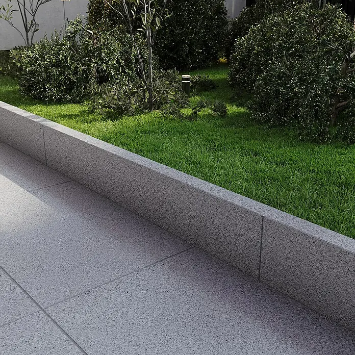 Rasenkante G 603 - 100 x 20 x 5 cm, Grau, Granit - Außenansicht mit Gartenanlage, die durch eine graue Steinmauer vom Gehweg getrennt ist. Die Bepflanzung besteht aus Büschen und Bäumen.