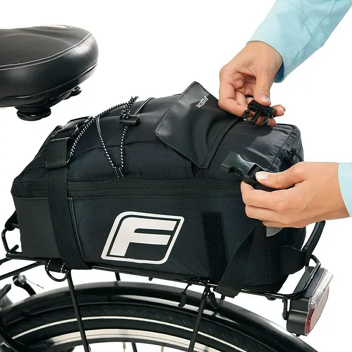 Fischer Gepäckträgertasche 2 in 1 - L x B x H: 18 x 16 x 32,5 cm - Nahaufnahme einer schwarzen Fahrradtasche mit weißem F-Logo, die an einem Gepäckträger befestigt und von Händen geschlossen wird.