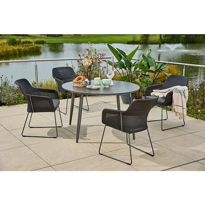 Sunfun Gartenmöbel-Set Solo - 5 -tlg., Aluminium/Sintered Stone/Polyester, Night-Grey - Ein stilvolles Outdoor-Set mit grauem Tisch und vier Stühlen auf einer Terrasse mit Blick auf einen See. Die Sitzgruppe lädt zum Verweilen ein.