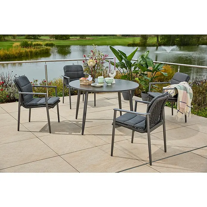 Sunfun Solo Gartentisch rund - Ø x H: 120 x 74 cm, Aluminium, Sintered Stone, Night-Grey - Ein Gartentisch-Set mit vier Stühlen auf einer Terrasse mit Blick auf einen See, perfekt für Mahlzeiten im Freien.