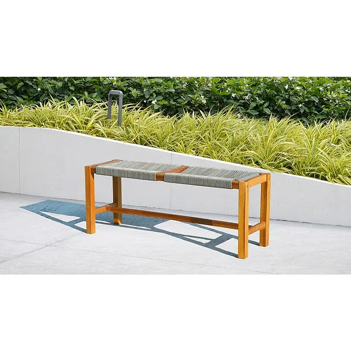 Banco de jardín Vilma - An x Pr x Al: 115 x 35 x 45 cm, Madera de acacia - Un banco de madera moderno con asiento tejido frente a plantas verdes y una pared blanca. Perfecto para espacios exteriores elegantes.