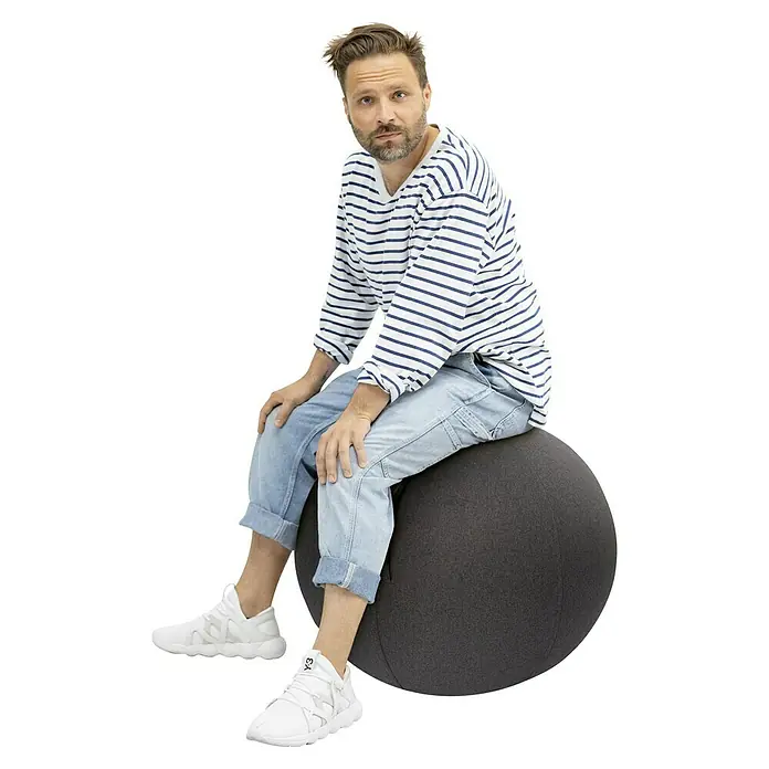 Sitting Ball Gymnastikball Felt - Anthrazit, Durchmesser: 65 cm, 100 % Polyester - Ein Mann sitzt auf einem grauen Gymnastikball, trägt ein gestreiftes Hemd und helle Jeans und weiße Turnschuhe.