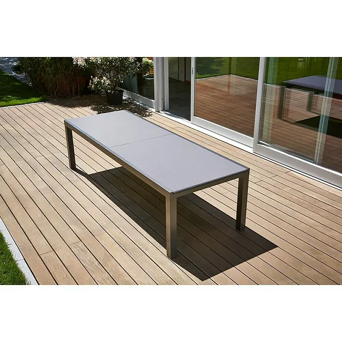 Sunfun Maja Gartentisch - L x B x H: 280 x 100 x 75 cm, Aluminium, Glas, Rechteckig, Night-Grey, Ausziehbar - Ein grauer Esstisch steht auf einer hölzernen Terrasse vor einer Glasschiebetür. Das moderne Design des Tisches passt gut in den Außenbereich.