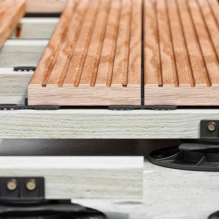 Langfit Abstandhalter - 4 mm, 100 Stk. - Nahaufnahme geriffelter Holz-Terrassendielen auf hellen Unterkonstruktionsbalken mit schwarzen runden Abstandshaltern, Teil eines erhöhten Decksystems.