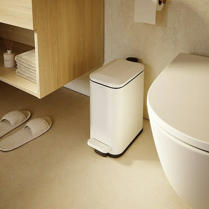 Cosmic Cubo de basura de baño Essentials Slim - 5 l, Metal, Blanco - Un elegante cubo de basura blanco con pedal y borde negro se encuentra en un baño moderno, junto a un inodoro y un mueble de madera.