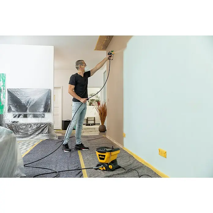 Wagner High Efficiency Airless Farbsprühsystem Control 150 M - 350 W, Förderleistung: 0,9 l/min - Ein Mann sprüht mit einer Wagner Farbspritzpistole eine Wand in einem Wohnzimmer, mit Abdeckmaterial zum Schutz des Bodens und der Möbel.