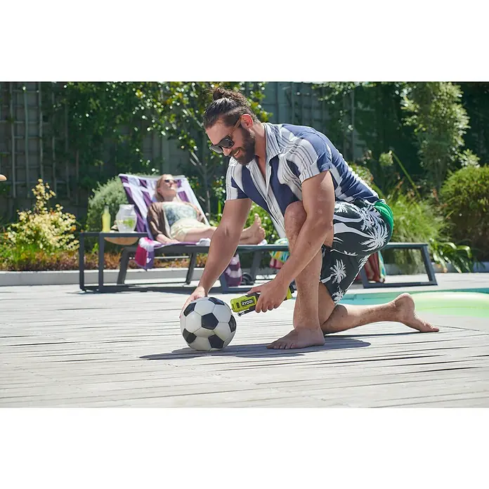 Ryobi 4V USB Akku-Kompressor Mini RI4-120G - 4 V, 1 Akku, 2 Ah, 6,9 bar - Ein Mann kniet neben einem Fußball und pumpt ihn mit einem gelben Ryobi-Inflator auf, während eine Frau sich im Hintergrund sonnt.