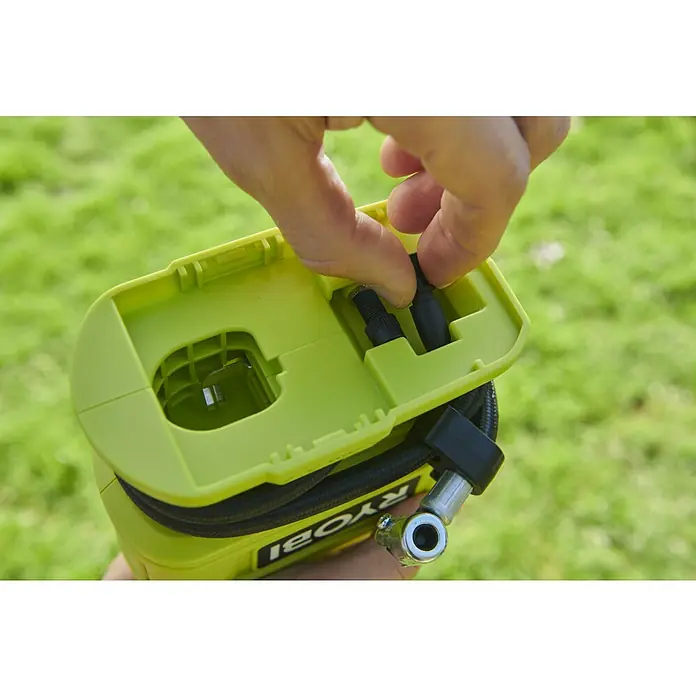 Ryobi ONE+ Akku-Kompressor RPI18-0 - 18 V, Ohne Akku, 10,3 bar - Detailaufnahme einer Person, die Zubehör in einem gelb-grünen Ryobi-Luftkompressor verstaut, vor einem verschwommenen grünen Hintergrund.