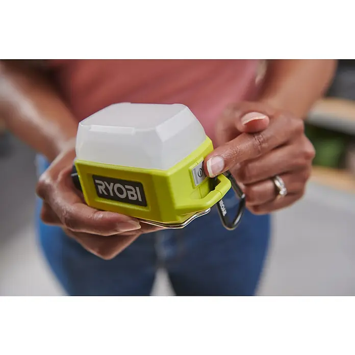 Ryobi 4V USB LED-Akkustrahler RLA4-0 - 300 lm - Nahaufnahme einer Person, die eine Ryobi-Campinglampe mit grünem Gehäuse und weißem Lampenschirm bedient, während sie blaue Jeans trägt.