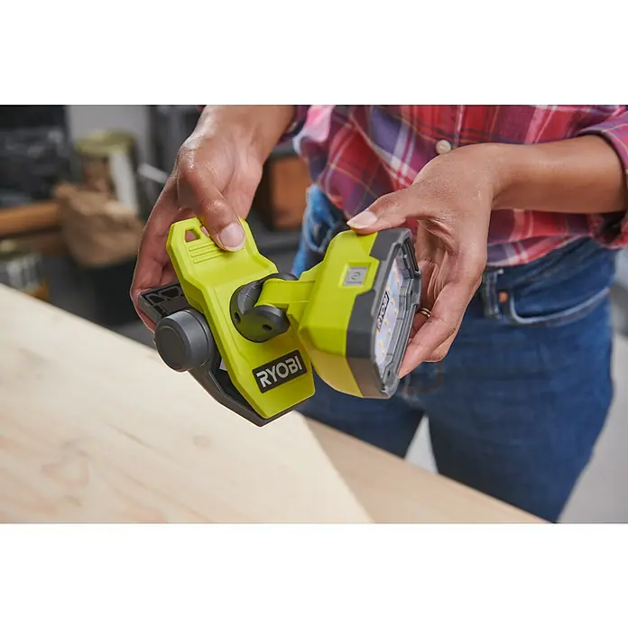 Ryobi 4V USB LED-Akkustrahler RLC4-0 - 1.000 lm - Nahaufnahme einer Person, die eine tragbare RYOBI-Arbeitsleuchte mit Klemme vor einem Holztisch hält.