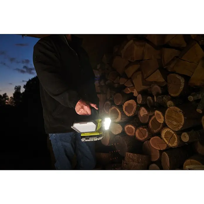 Ryobi ONE+ LED-Akkustrahler R18L360-0 - 3.500 lm, Akkuspannung: 18 V - Eine Person beleuchtet mit einer Akku-Arbeitsleuchte Holzscheite im Dunkeln. Die Lampe ist von Ryobi und scheint sehr hell zu sein.