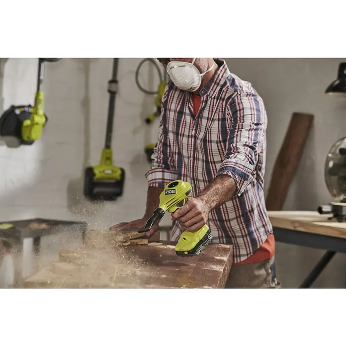 Ryobi ONE+ Akku-Luftpumpe RVI18-0 - 18 V, Ohne Akku, Max. Druck: 0,034 bar - Ein Mann verwendet einen gelben Ryobi-Akku-Gebläse, um Holzstaub von einem Stück Holz zu entfernen. Er trägt eine Staubmaske.