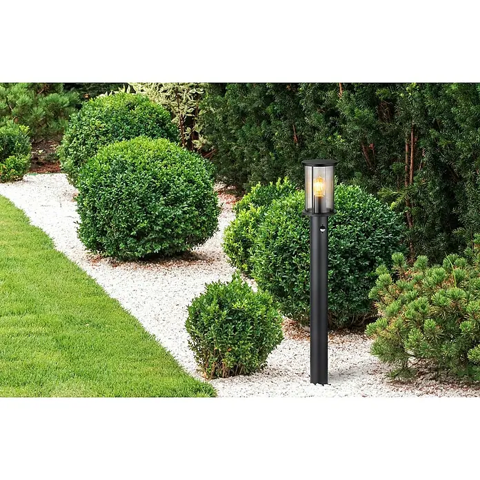 Globo Außenwandleuchte Gracey60 W, Anthrazit, IP54, Integrierter Sensor für automatisches Schalten - Gartenlampe aus dunklem Metall zwischen grünen Büschen auf einem Kiesweg. Die Lampe strahlt ein warmes Licht aus.