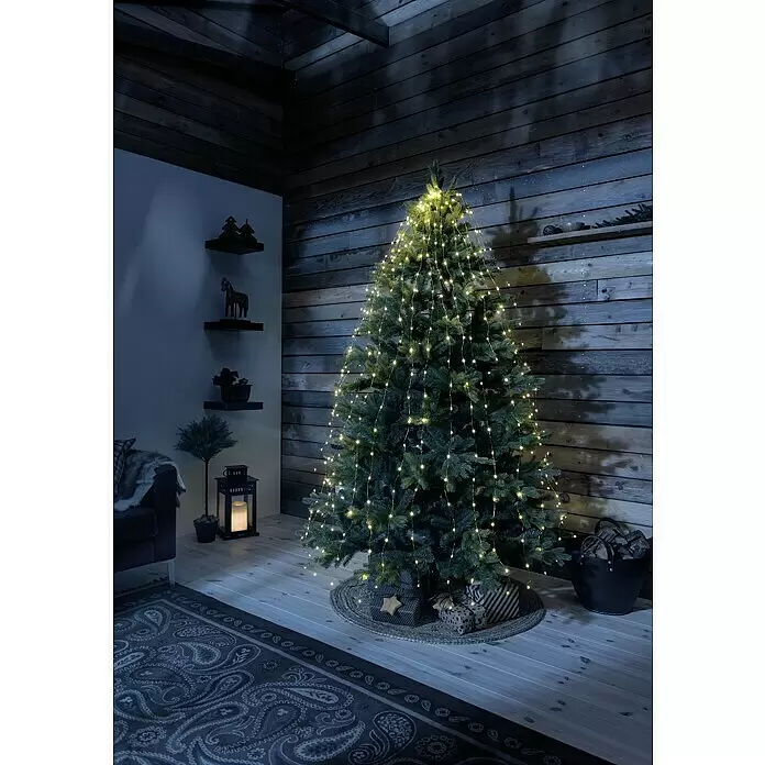 Konstsmide LED-Lichterkette Baummantel - Innen, Lichtfarbe: Bernstein, Anzahl Leuchtmittel: 180 Stk., Netzbetrieben - Weihnachtsbaum beleuchtet mit warmweißen Lichterketten in einem gemütlichen, schummrigen Raum.