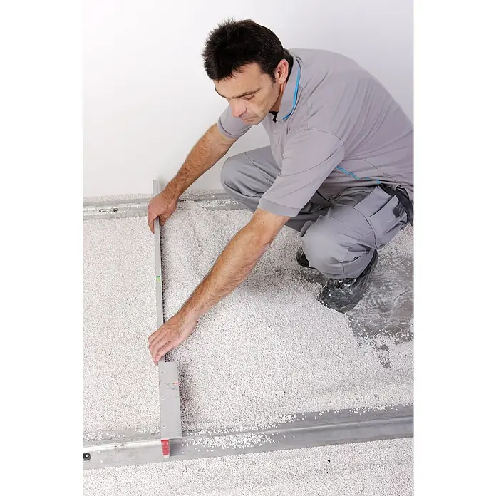 Knauf Perlite Ausgleichsschüttung Staubex® - 100 l - Ein Handwerker, der mit einer Wasserwaage einen Boden aus weißen Kieselsteinen nivelliert, um ein ebenes Fundament zu schaffen.