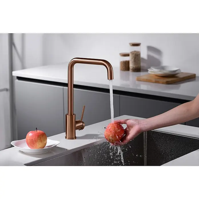 Imex Grifo de cocina Loira - Oro rosa - Primer plano de un grifo de cocina moderno en tonos cobrizos con una persona lavando una manzana en el fregadero bajo el agua corriente.