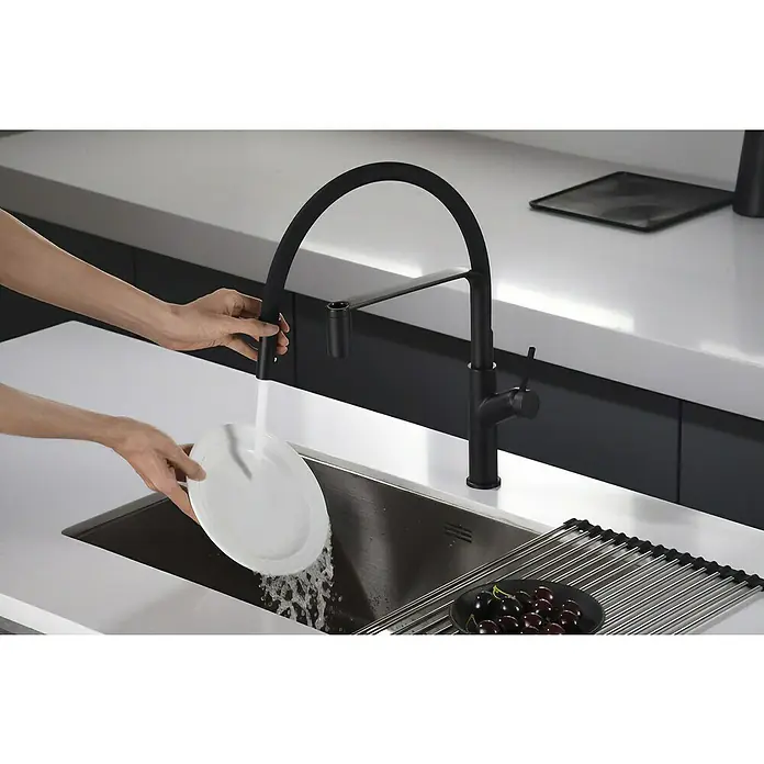 Imex Grifo de cocina flexible Sena - Negro - Una persona lava un plato bajo un grifo negro moderno en una cocina elegante con encimeras blancas.