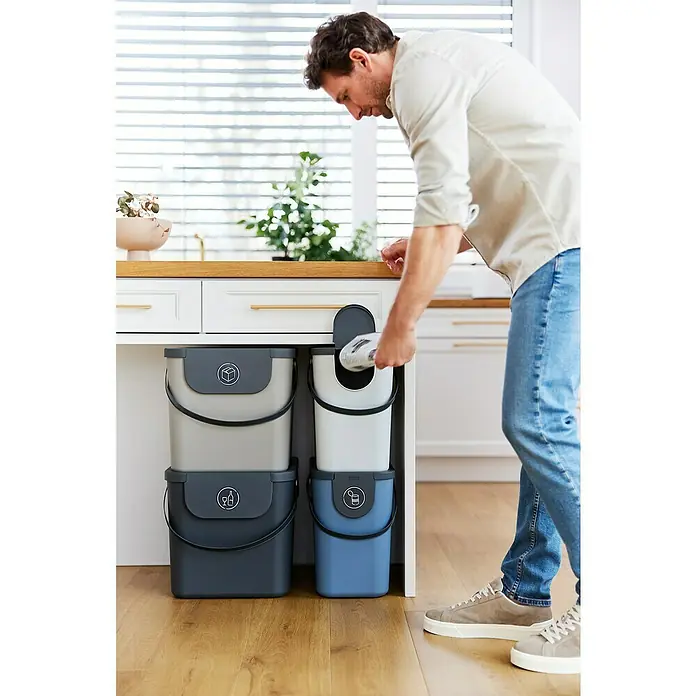 Rotho Albula Mülltrennsystem - 40 l, Kunststoff, Cappuccino - Mann wirft Abfall in ein Sortiersystem mit grauen, weißen und blauen Behältern unter einer Kücheninsel.