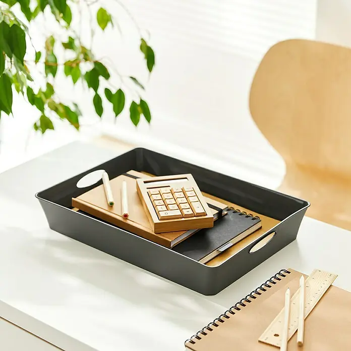Rotho Aufbewahrungsbox Living - L x B x H: 37,5 x 27 x 6 cm, Kunststoff, Anthrazit - Ein grauer Ablagekorb mit Büromaterialien wie Notizbüchern, Stiften und einem Taschenrechner auf einem Schreibtisch, der für Ordnung sorgt.