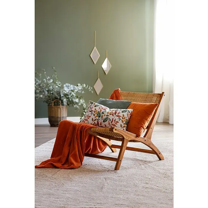 Cojín Pana - 45 x 45 cm, Poliéster, Verde, Cuadrado - Rincón de salón decorativo con una silla de mimbre con cojines y una manta naranja sobre una alfombra clara, con pared verde al fondo.