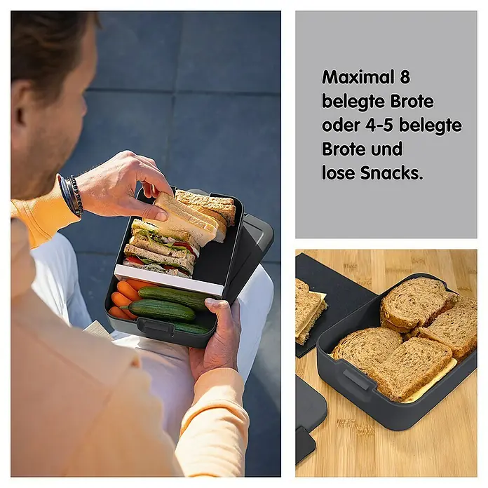 Sunware Sigma home Lunchbox Food to go - L x B x H: 24 x 17 x 6,5 cm, Anthrazit - Ein Mann befüllt eine graue Brotdose mit mehreren Fächern, die Sandwiches, Gurken und Karotten enthält.
