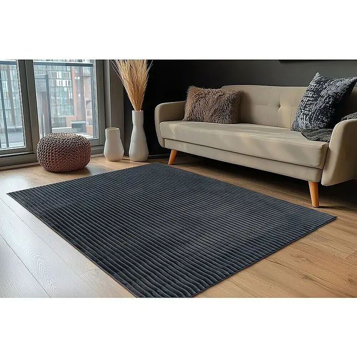 Hochflorteppich Plush - Graphite, 230 x 160 cm, 100 % Polyester - Ein grauer Teppich mit einem gerippten Design liegt auf einem Holzboden, ergänzt durch ein Sofa und dekorative Elemente.