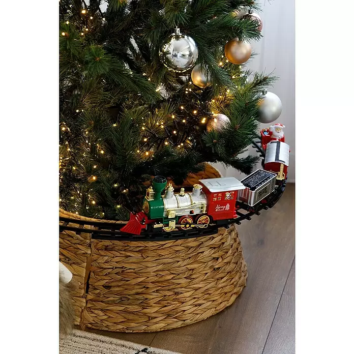 Elemento decorativo Tren LED - Colores surtidos, Plástico, 1 ud. - Un tren de juguete festivo verde, rojo, plata y oro que rodea la base de un árbol de Navidad decorado en una cesta tejida.