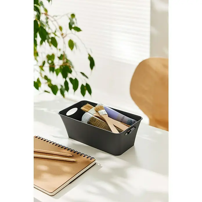 Rotho Aufbewahrungsbox Living - L x B x H: 25,2 x 13,4 x 9 cm, Kunststoff, Anthrazit - Ein dunkler, rechteckiger Aufbewahrungskorb mit Griffen, gefüllt mit Pinseln und Farben, steht auf einem Schreibtisch neben einem Notizbuch.