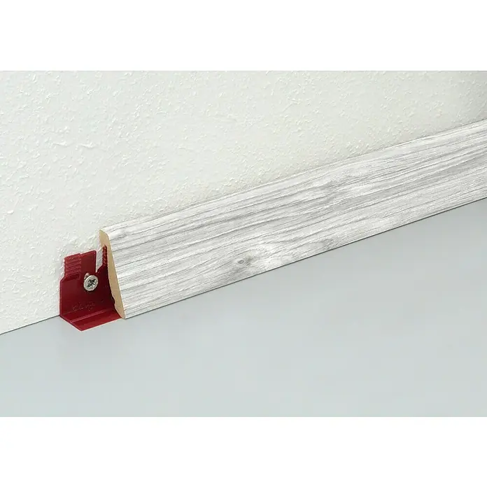 Profiles and more Sockelleiste Tarronga FU147H - 2.400 x 18,5 x 38,5 mm, MDF - Nahaufnahme einer Sockelleiste in Holzoptik, befestigt mit einem roten Clip an einer weißen Wand, die den Boden berührt.