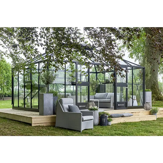 Juliana Gewächshaus Grand Oase - Außenmaß inkl. Dachüberstand (B x T): 368 x 511 cm, 3 mm, Anthrazit/Schwarz - Ein Juliana Gewächshaus mit schwarzem Rahmen auf einer Holzplattform, umgeben von Grün und Gartenmöbeln.