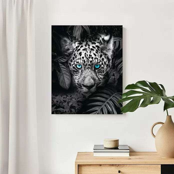 Reinders Decopanel - Blue Eyed Leopard, B x H: 40 x 50 cm - Leinwandbild mit einem schwarz-weißen Leoparden mit leuchtend blauen Augen im Dschungel, an einer Wand über einem Holztisch.
