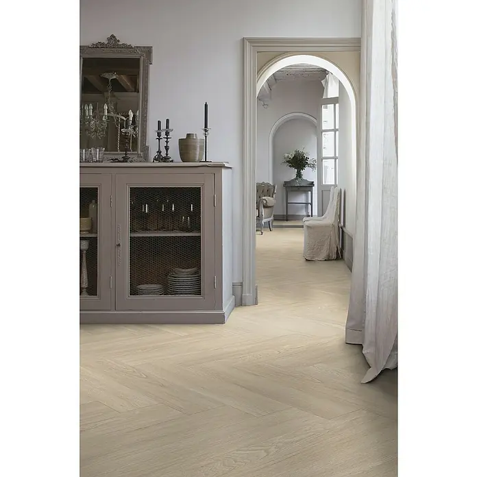Gerflor Vinylboden Rigid Acoustic - Tulum Light, Fischgrät, 750 x 125 x 6 mm - Elegantes Interieur mit hellen Holzböden im Fischgrätmuster, einem grauen Sideboard mit Spiegel und Blick durch einen Torbogen in ein weiteres Zimmer.