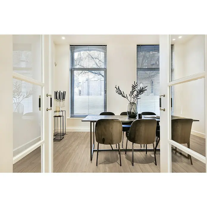 Gerflor Vinylbodenmuster Rigid Acoustic Serra Beige - 250 x 229 x 5,4 mm, Serra Beige - Elegantes Esszimmer mit Holztisch, Polsterstühlen und weißen Glastüren, beleuchtet durch Fenster mit Jalousien.