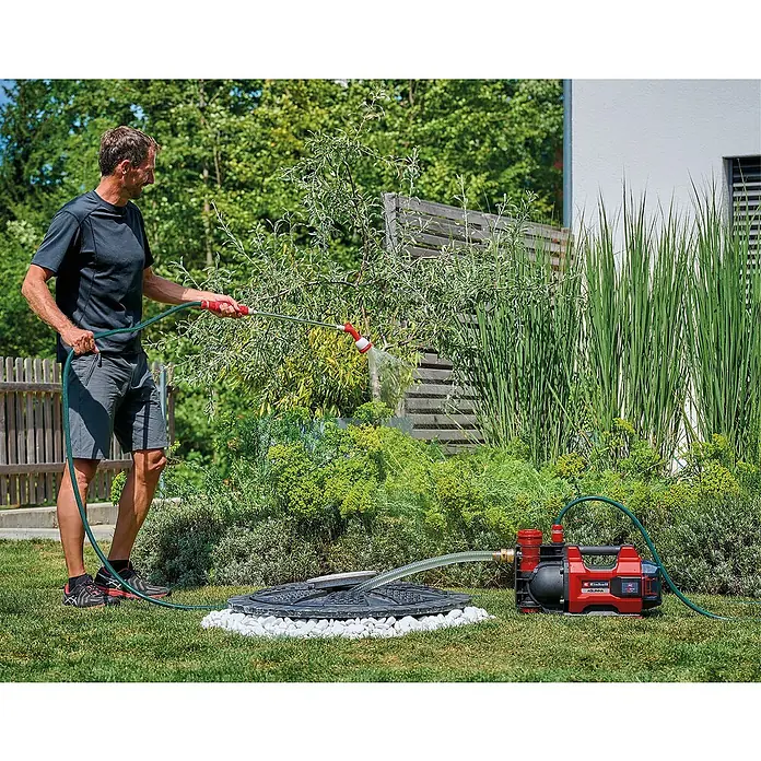 Einhell Power X-Change 18V Akku-Gartenpumpe Aquinna 36/38 F LED Automatic - 18 V, Li-Ionen, Ohne Akku, Max. Fördermenge: 3.800 l/h - Ein Mann bewässert seinen Garten mit einer roten Einhell Gartenpumpe, die auf einer Rasenfläche neben einem Brunnen steht.