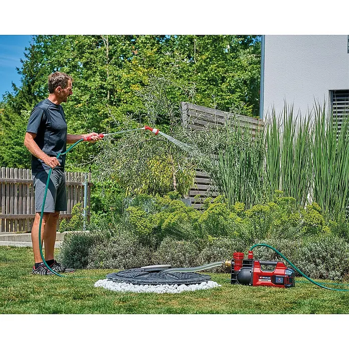 Einhell Power X-Change 18V Akku-Gartenpumpe Aquinna 36/38 F LED - 18 V, Li-Ionen, Ohne Akku, Max. Fördermenge: 3.800 l/h - Ein Mann bewässert Pflanzen mit einem Einhell Hauswasserwerk. Das Gerät ist rot und schwarz und steht auf dem Rasen.