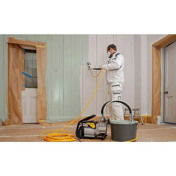 Wagner High Efficiency Airless Farbsprühsystem SuperFinish 21 Pro HEA - 900 W, Förderleistung: 2,1 l/min - Ein Handwerker benutzt eine Wagner Farbspritzpistole, um eine Wand zu streichen, wobei er Schutzkleidung trägt und die Umgebung abgedeckt ist.