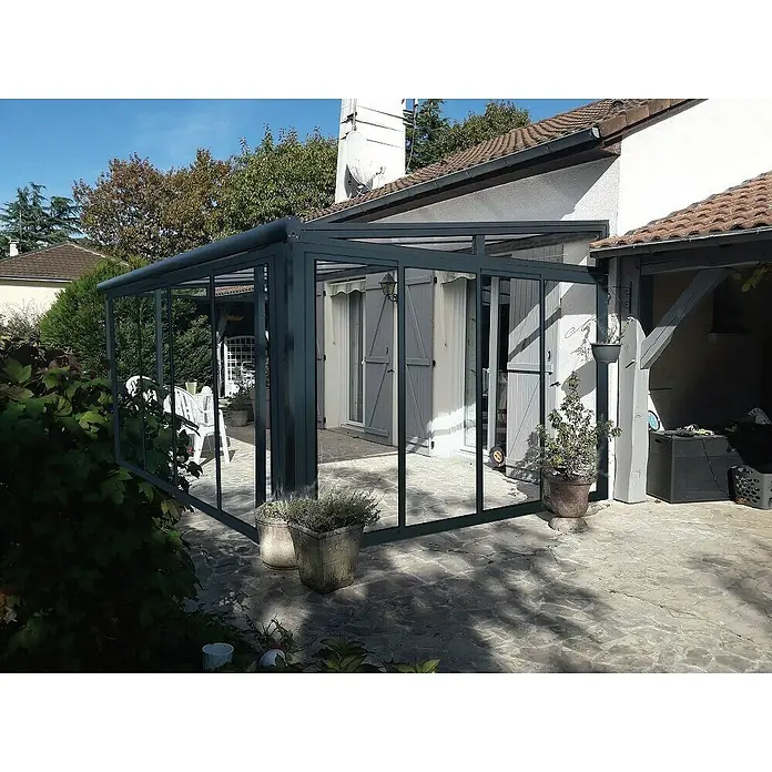 Wismar-NFI Wintergarten - 404 x 303,3 cm, Pultdach, Polycarbonat, Anthrazitgrau, Milchig Weiß - Nahaufnahme einer modernen, grauen Glasveranda, die an ein Haus angebaut ist und einen hellen, einladenden Außenbereich bietet.