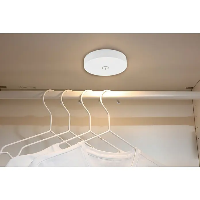 Tween Light Akku-Unterbauleuchte - Weiß, Mehrfarbig - Diese Nahaufnahme zeigt eine runde, weiße Schrankleuchte mit einem Power-Symbol an einer Schrankdecke über einer Kleiderstange mit Kleiderbügeln.