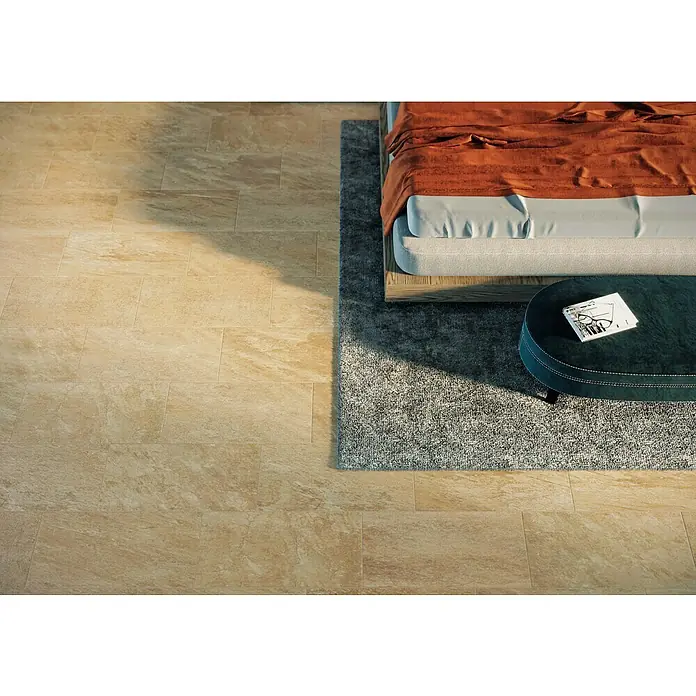 Atlas Concorde Solution Regent Feinsteinzeugfliese Gold - 30 x 60 cm, Gold, Matt - Beige Fliesen zieren den Boden neben einem Bett mit orangefarbener Decke und einem dunkelgrünen Hocker mit Buch.