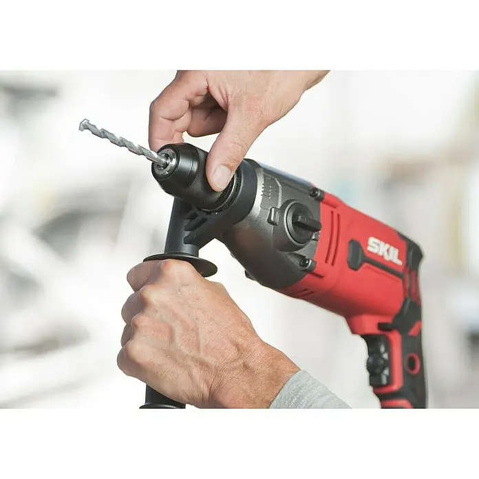 Skil Schlagbohrmaschine 6745 GA - 1.050 W, Schnellspannbohrfutter - Nahaufnahme einer SKIL-Bohrmaschine, die von Händen gehalten wird, während ein Bohrer eingesetzt wird, was ihre Benutzerfreundlichkeit hervorhebt.