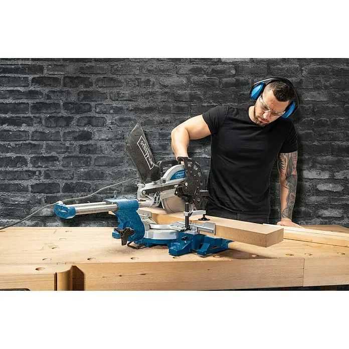Scheppach Tronzadora HM 254 - 2.000 W, Diámetro hoja de sierra: 255 mm, 4.500 r.p.m. - Un hombre utilizando una sierra ingletadora Scheppach para cortar madera, con gafas de seguridad y auriculares.