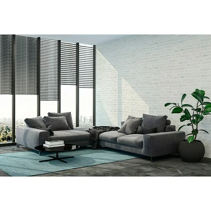 A.S. Création New Walls Vliestapete Stein - Weiß, Steinoptik, 10,05 x 0,53 m - Graues Ecksofa mit Kissen, Couchtisch und Pflanze in einem hellen Raum mit Jalousien und Ziegelwand.