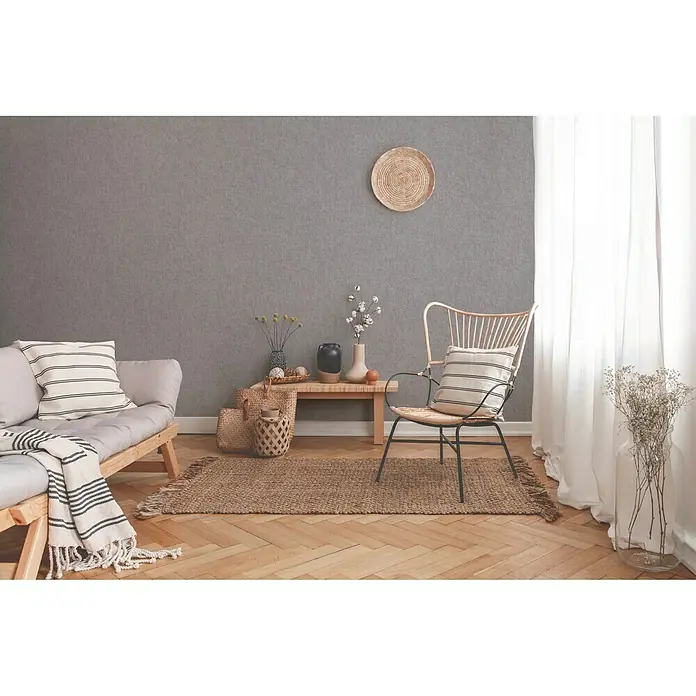 A.S. Création New Walls Vliestapete - Taupe, Uni, 10,05 x 0,53 m - Wohnzimmer-Interieur mit Rattan-Stuhl, Teppich und Couch in einem natürlich eingerichteten Raum mit Holzboden und grauer Wand.
