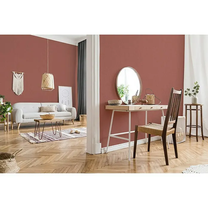 A.S. Création New Walls Vliestapete Textil - Rot, Uni, 10,05 x 0,53 m - Einladendes Wohnzimmer mit terrakottafarbenen Wänden, einem hellen Sofa und einem Holztisch. Ein Spiegel steht auf einer Kommode mit einem Stuhl.
