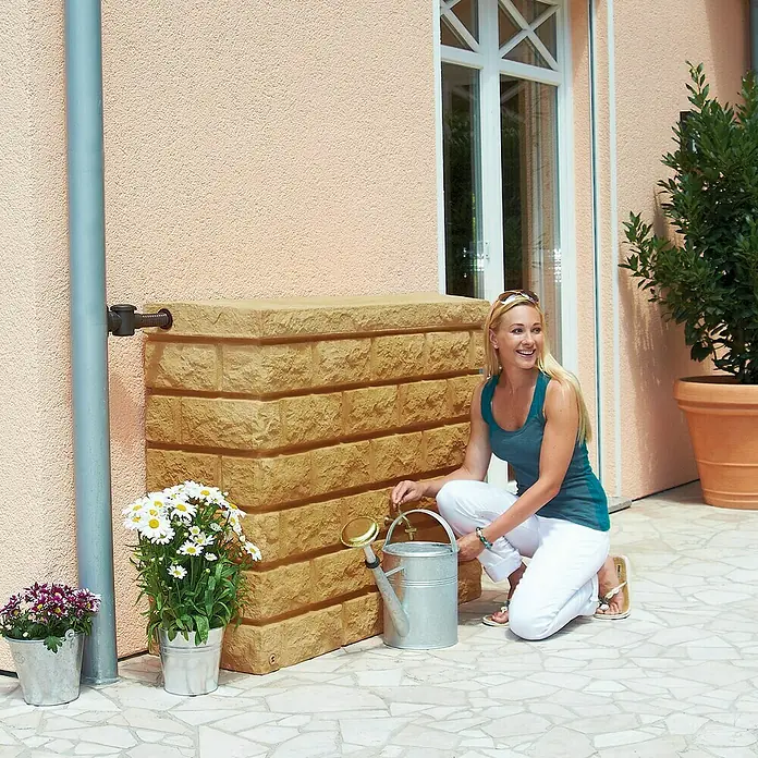 Garantia Exklusive Line Regenspeicher Rocky - 400 l, Sandsteinfarben - Eine Frau befüllt eine Gießkanne an einem Sandfarbenen Wasserbehälter mit Steinoptik, der an einer Hauswand steht.