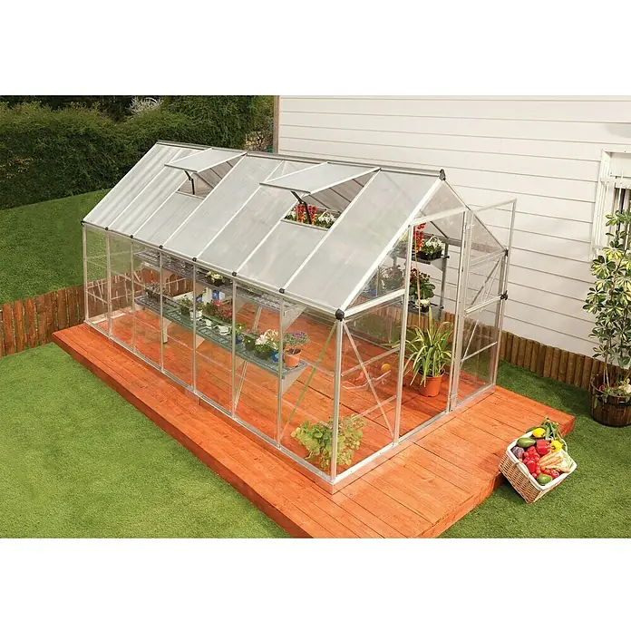 Palram – Canopia Gewächshaus Hybrid - Außenmaß inkl. Dachüberstand (B x T): 185 x 426 cm, 0,7 mm, Silber - Das Gewächshaus mit Glaswänden und Pflanzen darin, auf einem Holzpodest im Garten. Ein Korb mit Gemüse steht daneben.