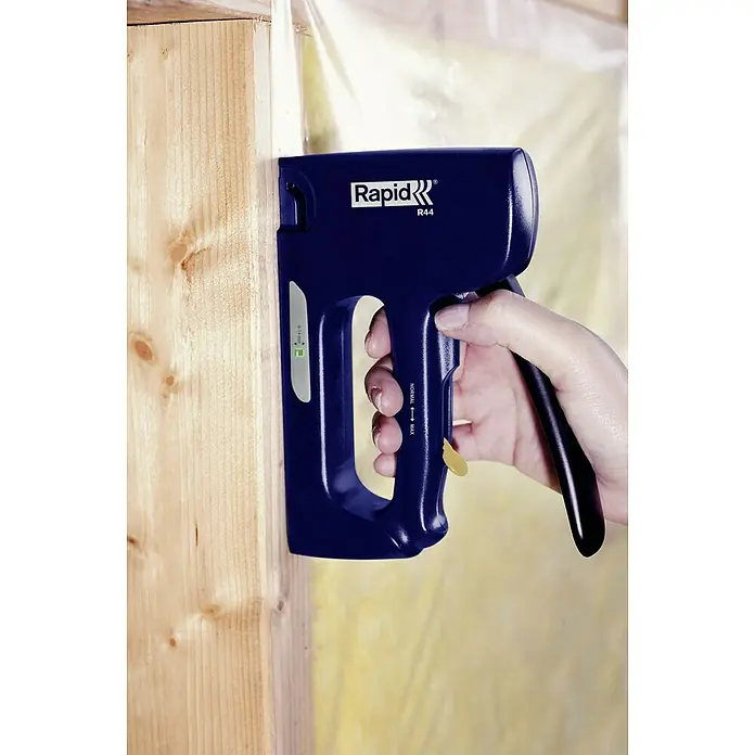 Rapid Handtacker Rapid R44 - Klammertiefe: 4 mm - 14 mm, Geeignet für: Flachdrahtklammern Nr. 140 - Nahaufnahme eines Rapid R44-Tackers, der zum Befestigen von Materialien an einer Holzwand verwendet wird, mit einer Hand, die den Griff betätigt.